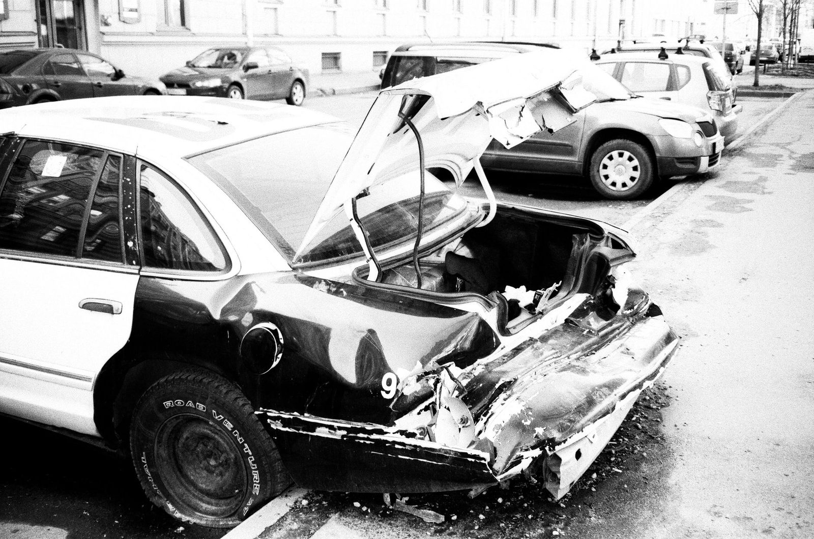 A black and white photo of a wrecked car with heavy front-end damage on an urban street.