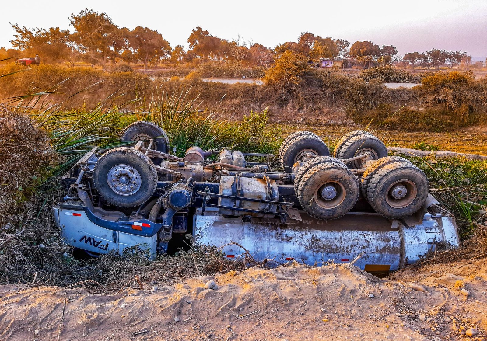 A large truck lies overturned on the shoulder of a rural dirt road in daylight.