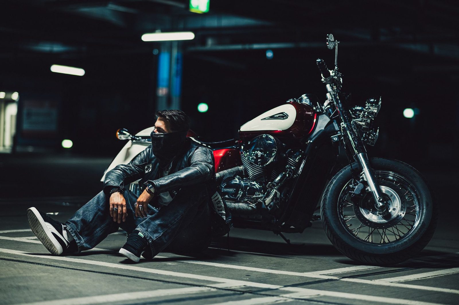 A man in a leather jacket sits beside a motorcycle in a dimly lit garage.