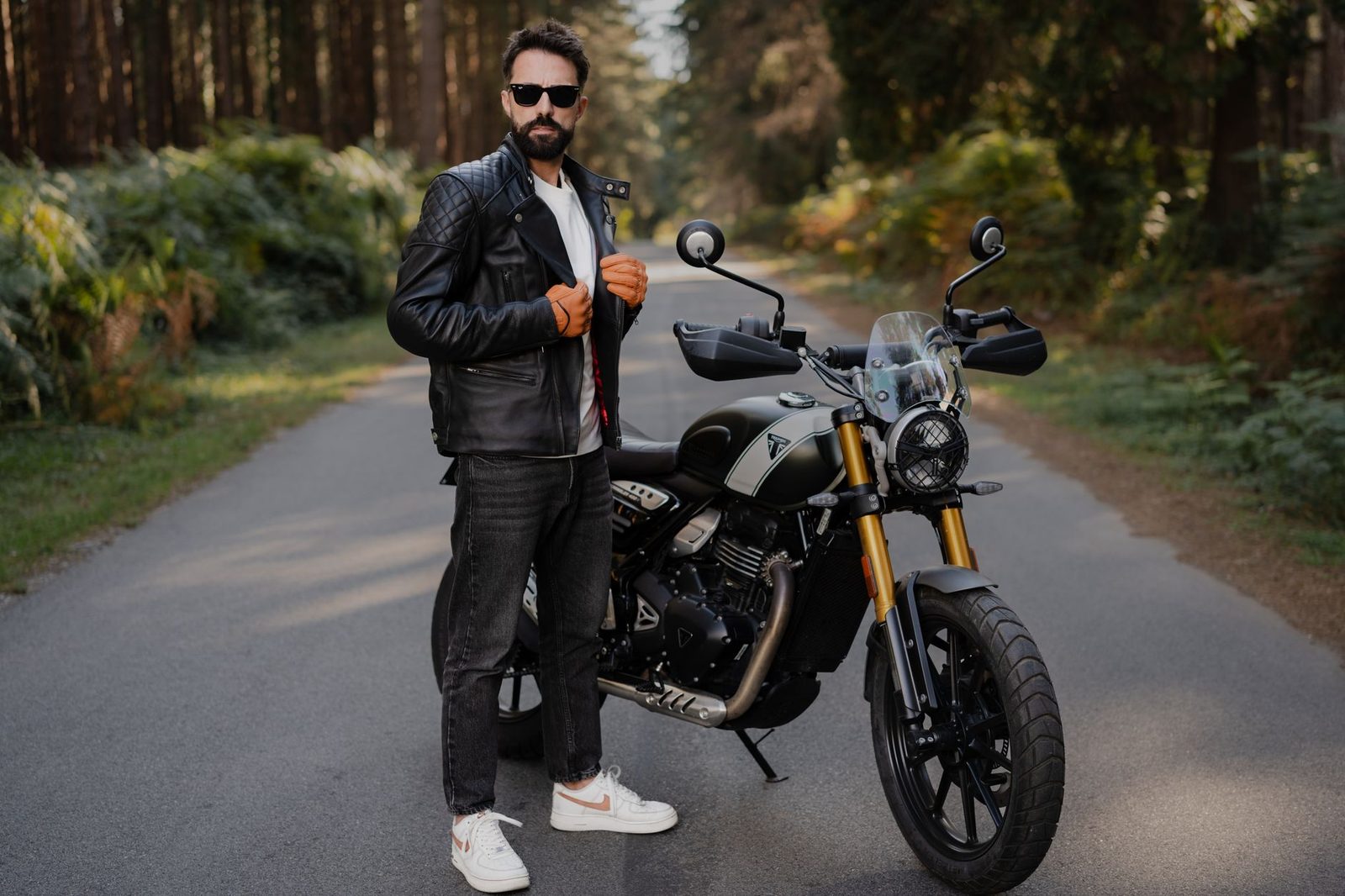 A man in stylish clothing poses beside a motorcycle on a road lined with forest.