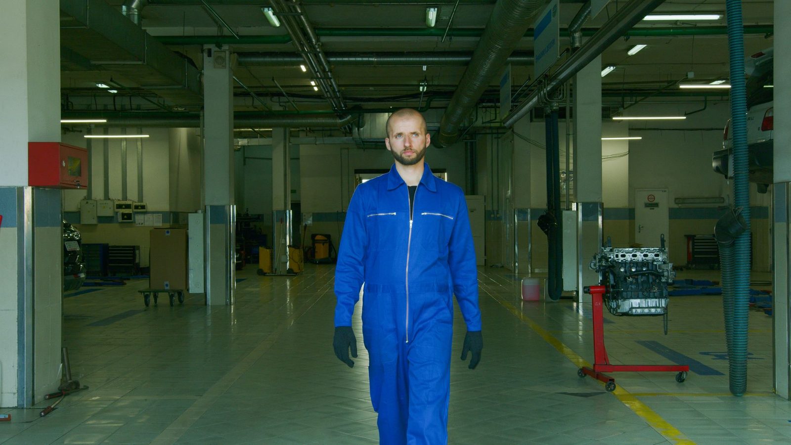 A mechanic in a blue jumpsuit walks confidently through a modern auto repair workshop.