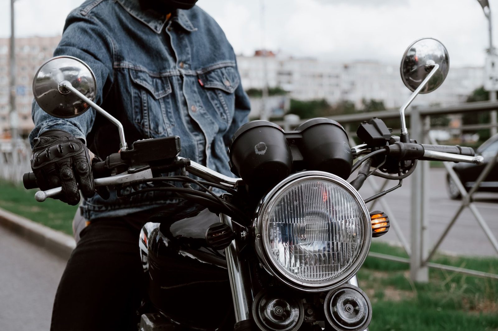 A motorcyclist in a denim jacket grips the handlebars on a city street.