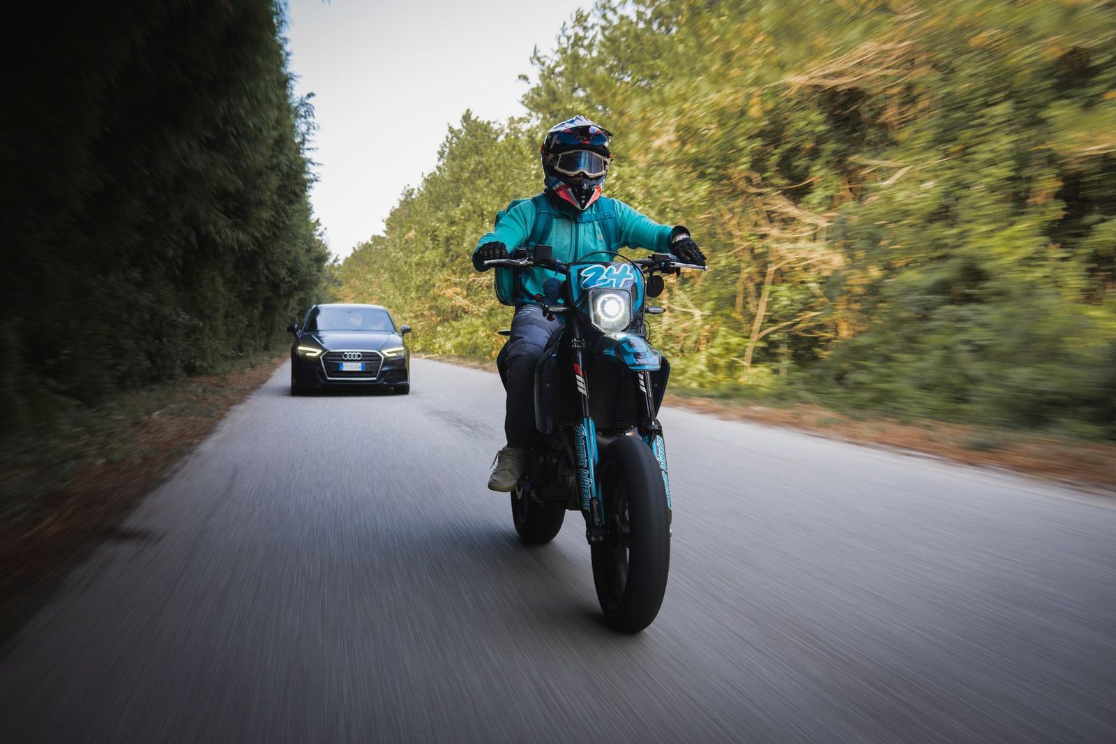 A motorcyclist rides past an Audi car on a rural road, conveying speed and motion.