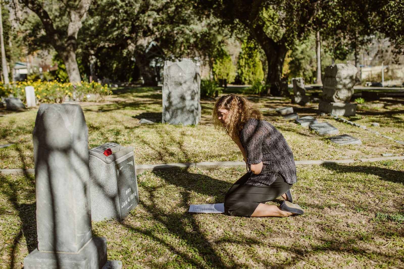 A woman kneels by a gravestone in a sunlit cemetery, showing grief.
