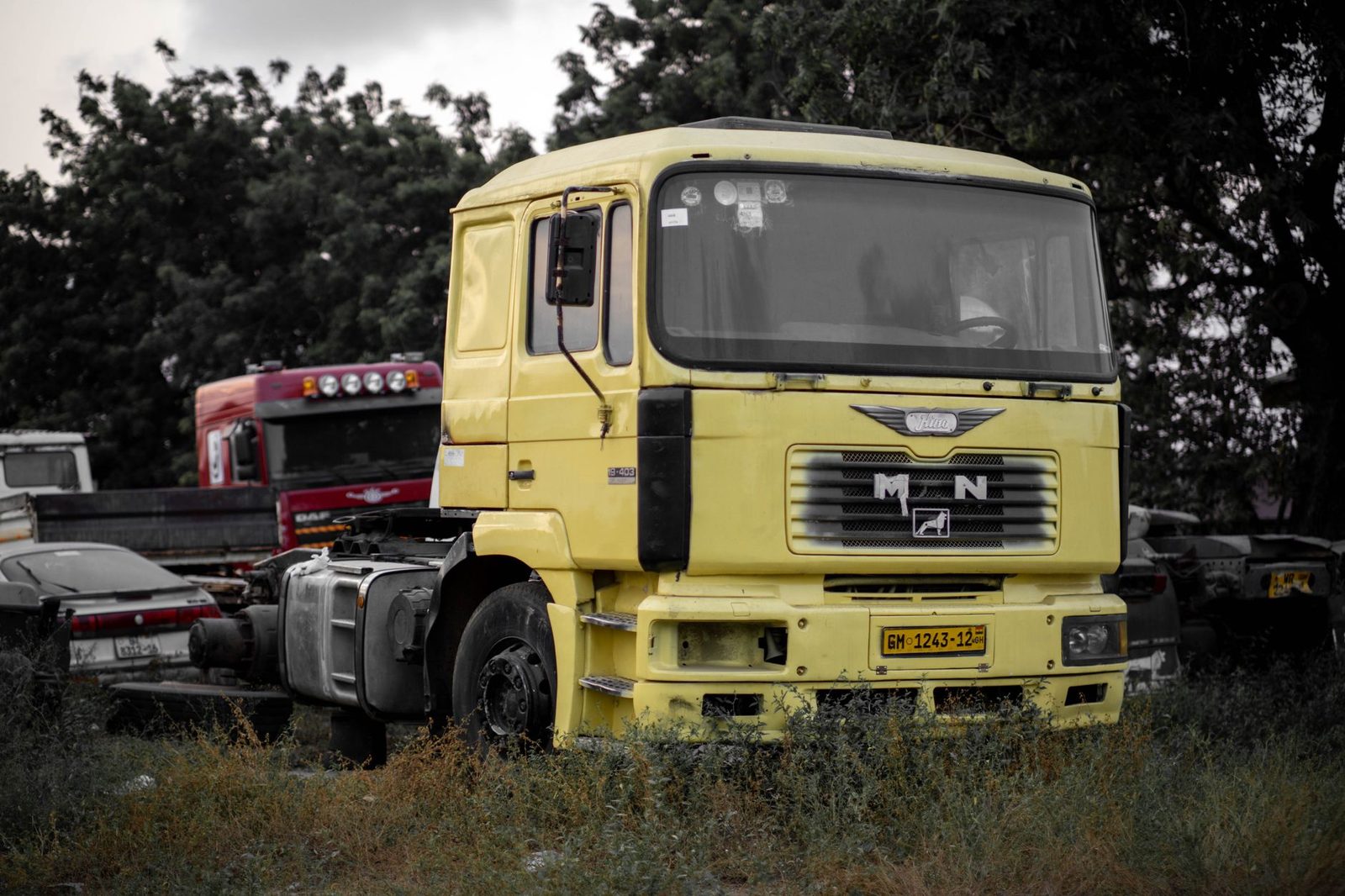 A yellow semi-truck cab parked in an outdoor lot among other vehicles and trees.