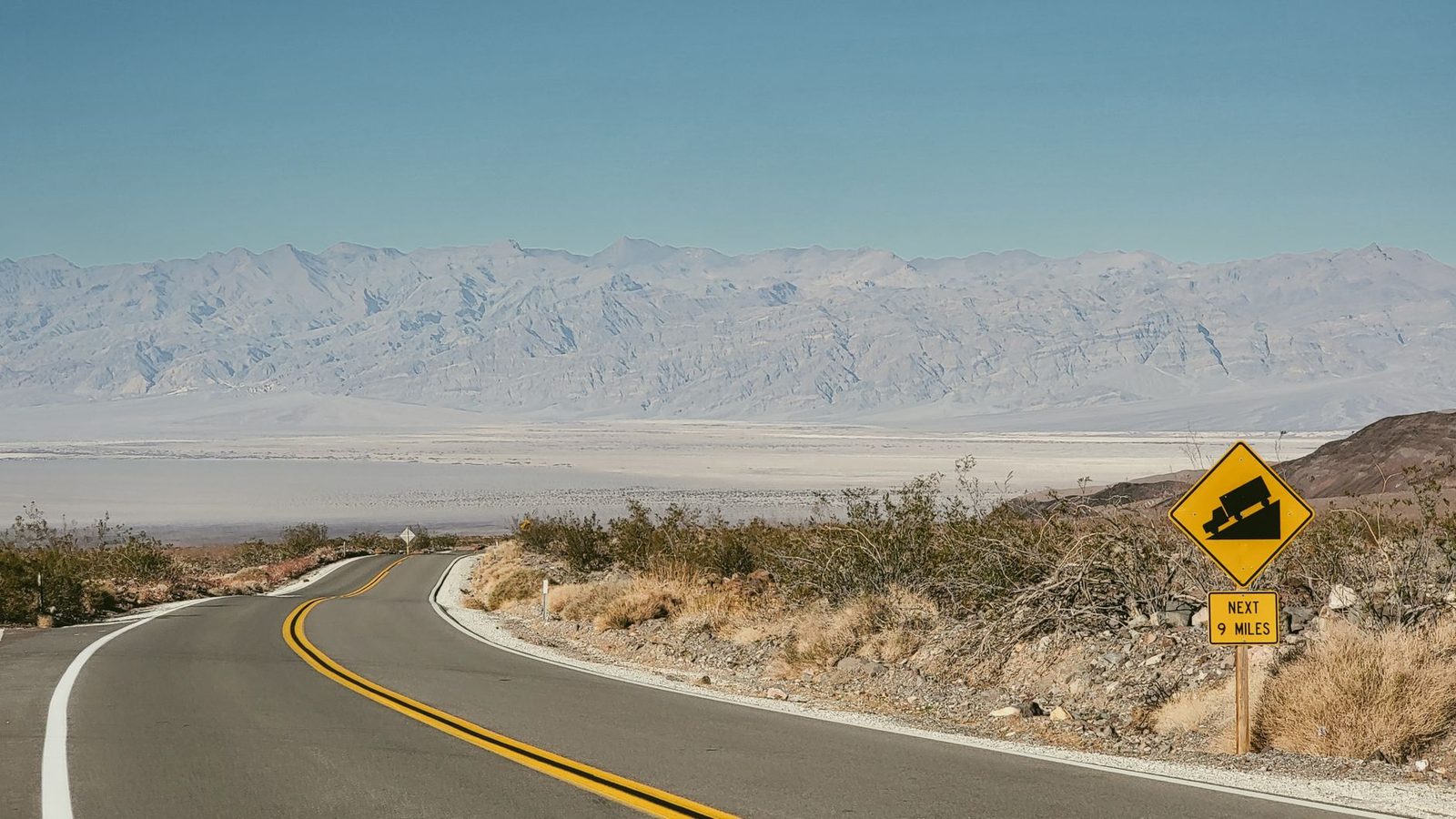 An empty road curves through an arid desert landscape toward distant mountains.