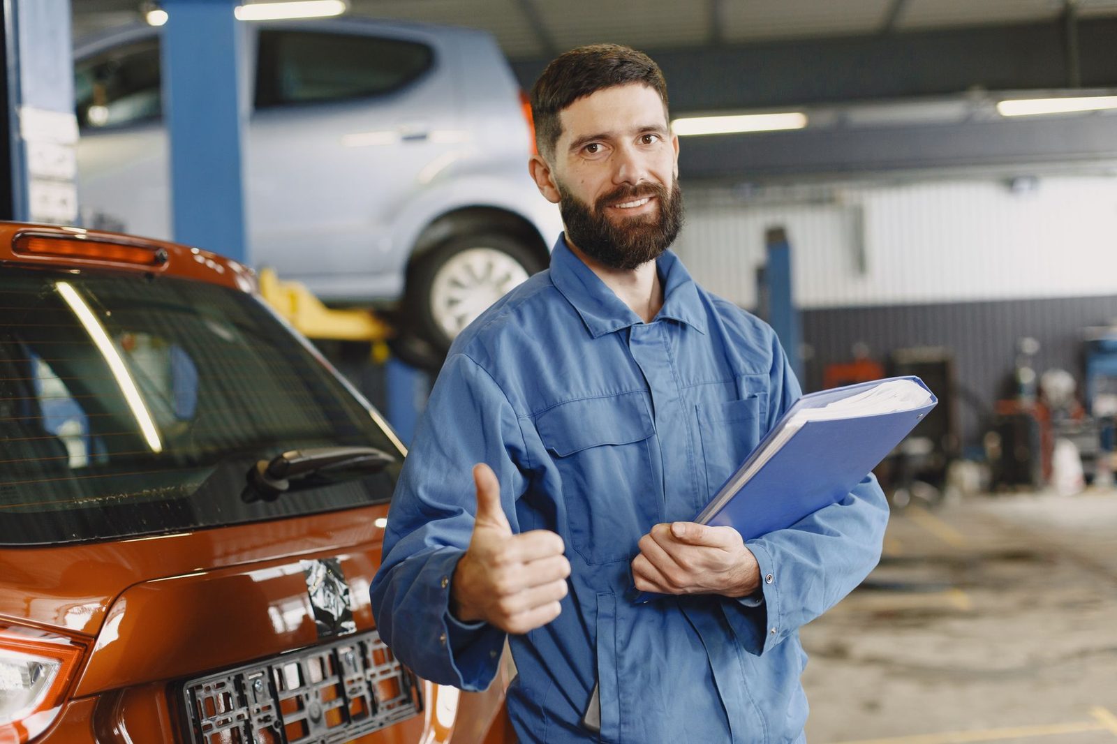 Auto mechanic in blue coveralls holding a clipboard and giving a thumbs up in a garage