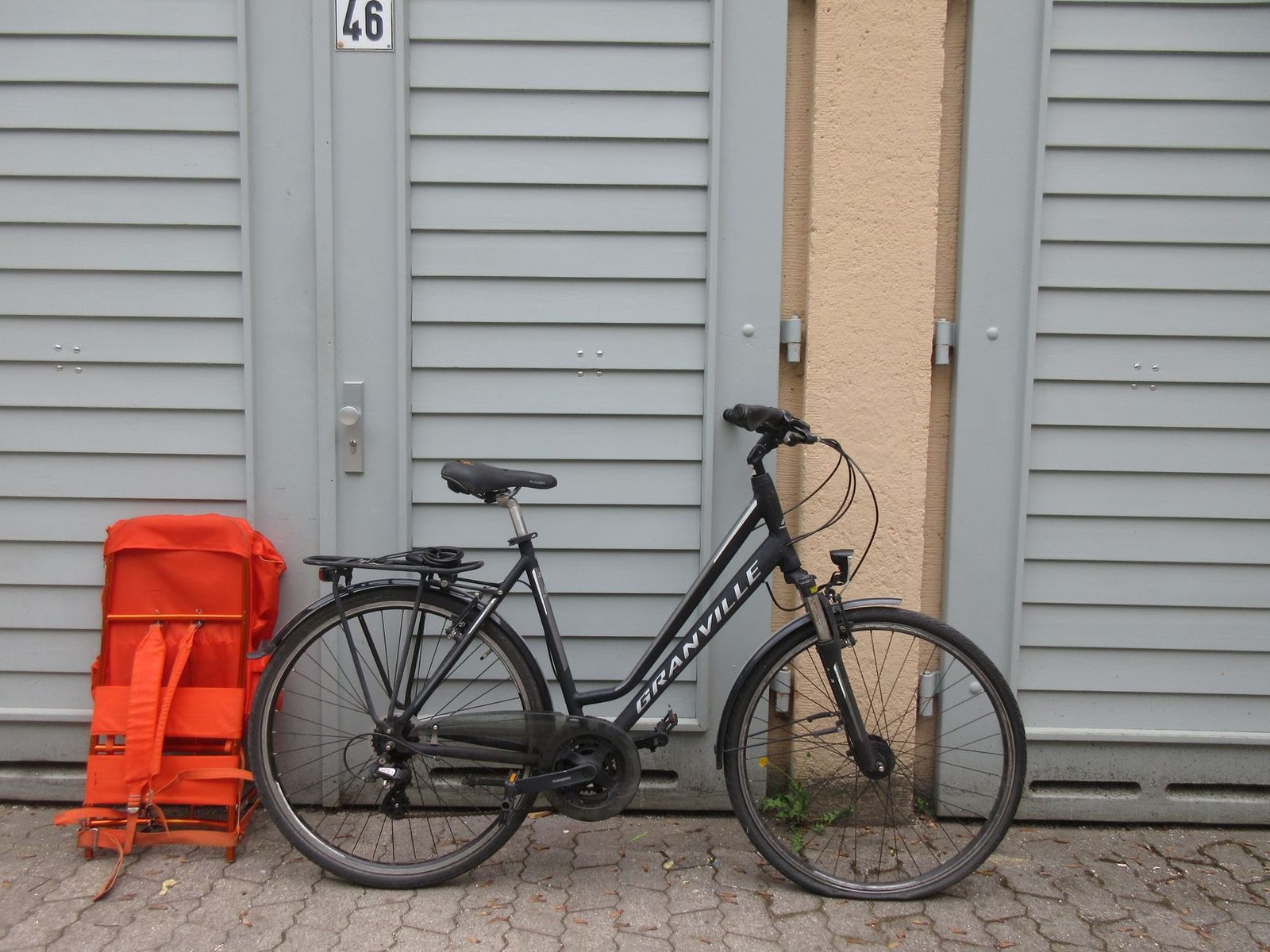 Black bicycle with a bright orange backpack leaning against a closed garage door on a paved city street.