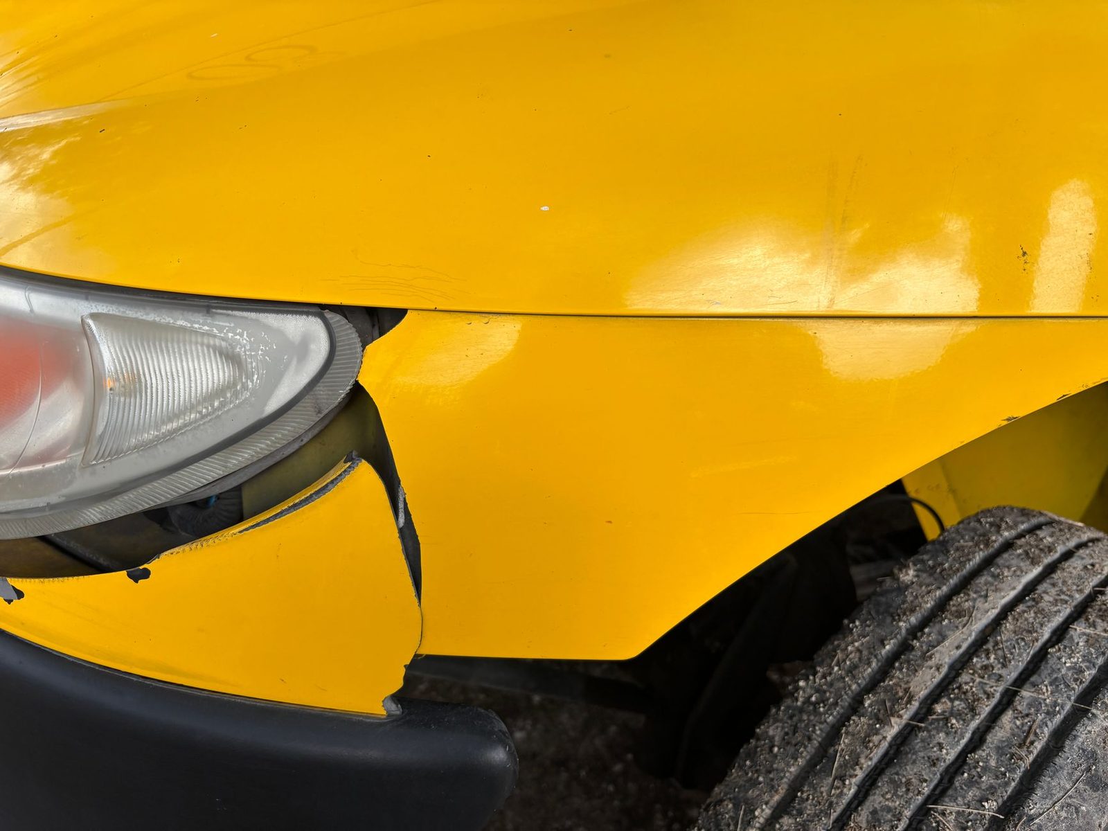 Close-up of a yellow car's dented front bumper and cracked headlight after a collision.