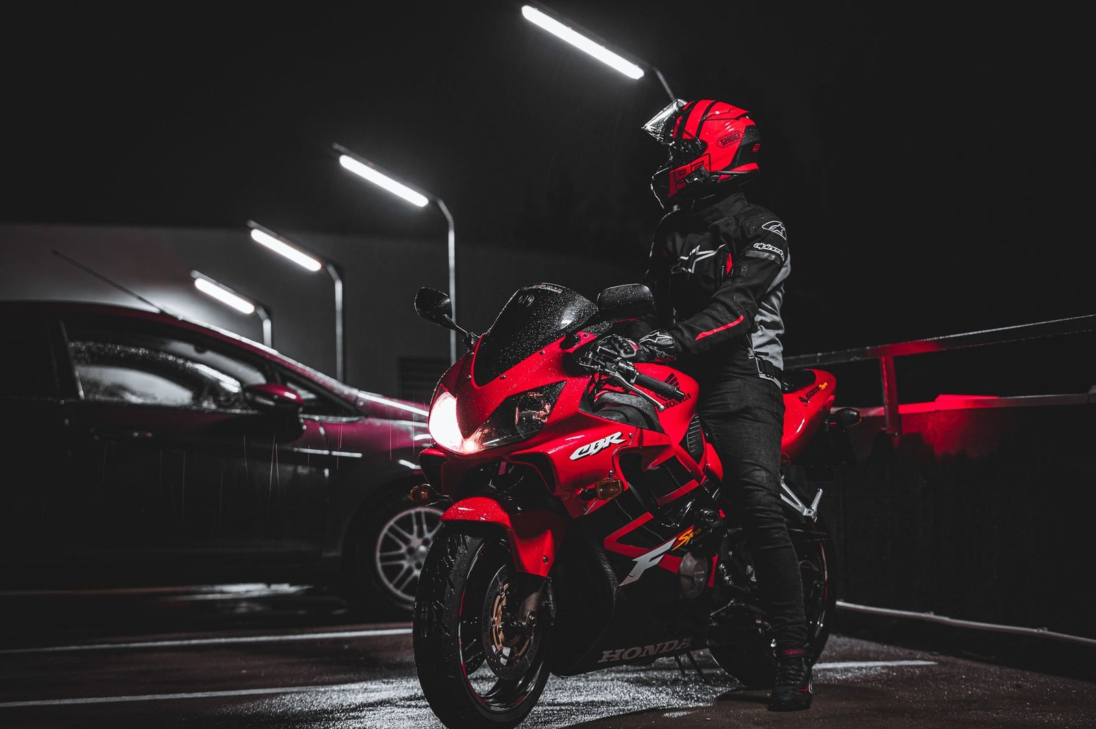 Motorcyclist in red riding gear stands beside a red sport motorcycle in a dimly lit parking area at night.