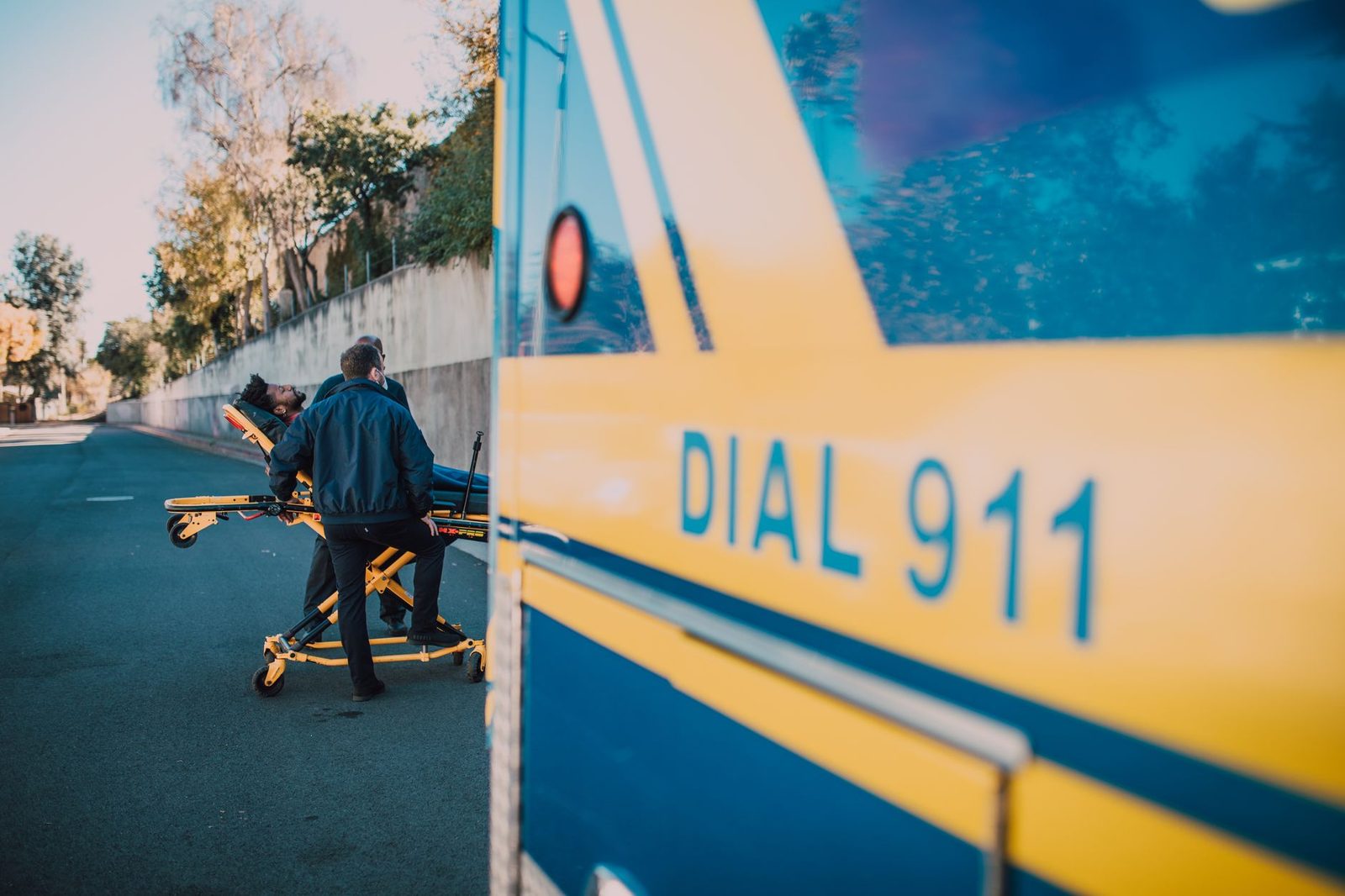 Paramedics help a patient on a stretcher beside an ambulance on a sunny day.