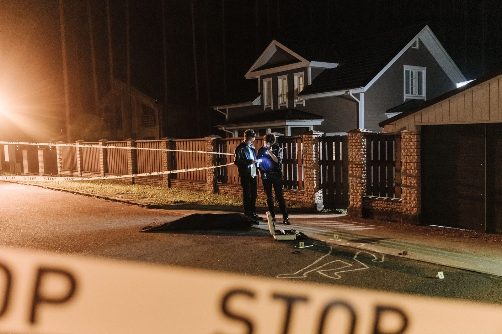 Police officers work at night outside a house, marking evidence beside yellow crime scene tape.