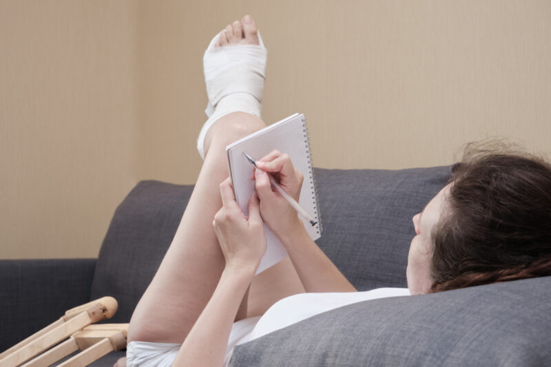 Woman in a plaster cast writing in notebook lying on a couch in the living room at home