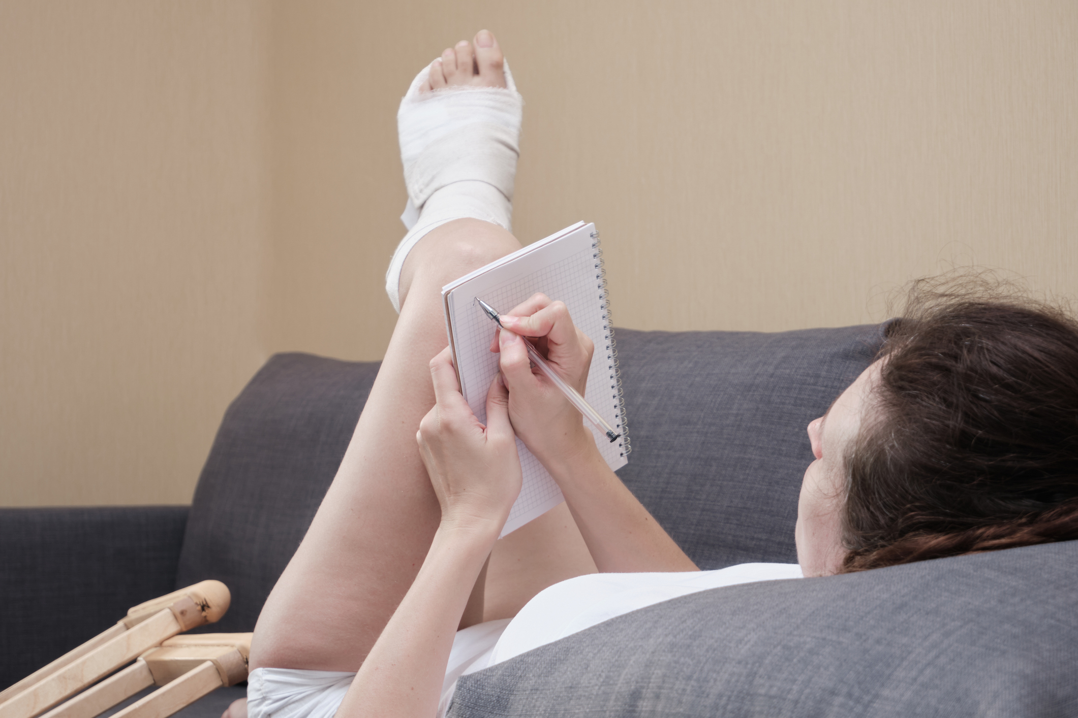Woman in a plaster cast writing in notebook lying on a couch in the living room at home